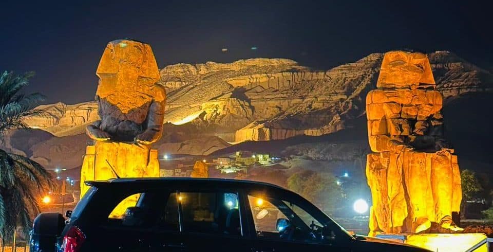 Black SUV parked at night in front of the illuminated Colossi of Memnon in Luxor, Egypt.